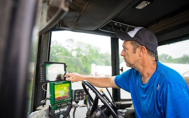 A man sits in the cab of a tractor. Two small computer display screens sit in front of him behind the steering wheel providing information to help him apply fertilizer to his crop.