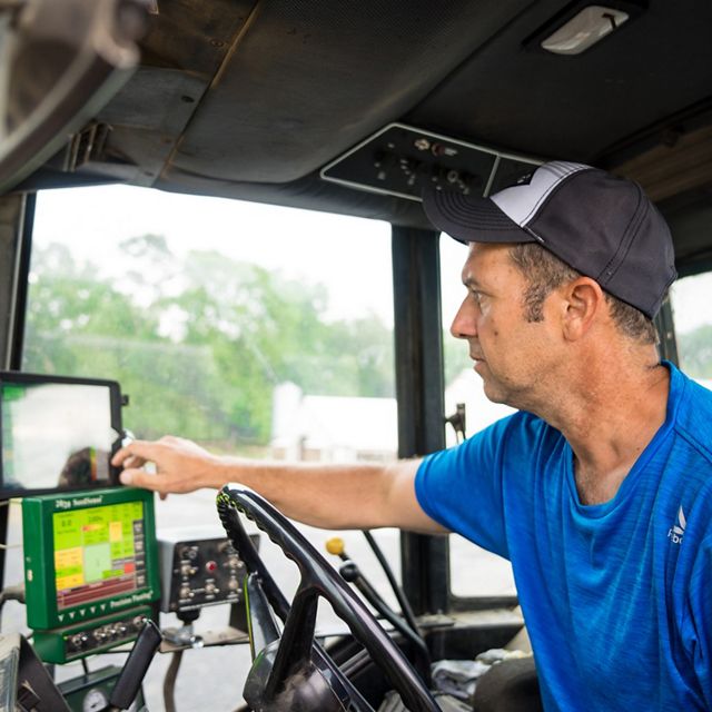 A man sits in the cab of a large tractor using small computer monitors to held determine the amount of fertilizer to apply to his field.