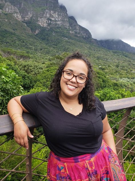Andrea van Wyk headshot. Her arm rests on the top of a fence. A vegetation filled valley extends behind her, meeting the rocky, steep face of a mountain. Thick white clouds hang low in the sky.