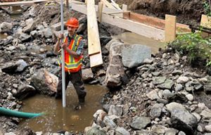 A river restoration expert wearing a hard hat and high‑visibility vest stands in a shallow stream, holding a measuring rod amid rocks and active construction.
