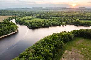 The sun rises over a wide river that splits into two branches, flowing past tree lined banks and open fields.