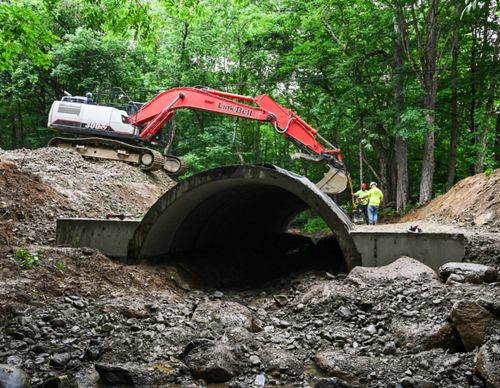 Construction workers and equipment in a forested area next to a large concrete structure.