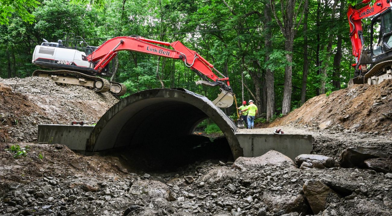Construction workers work in a forested area with a large, concrete structure perched over a stream.