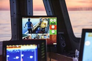 A monitor on a research vessel shows live video feeds of scientists working on deck during an aquatic research project. Through the surrounding windows, a pink and orange sunset reflects off calm water.