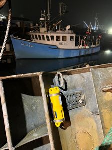 At a harbor at night, a weathered metal trap fills the foreground. Attached to the trap is a bright yellow cylindrical sensor secured with metal bands. A metal identification plate on the trap shows the markings “01033” and “WFS125.” In the background, a blue fishing vessel named Emma Rae is tied to the dock, reflected in the dark, still water under artificial lights.