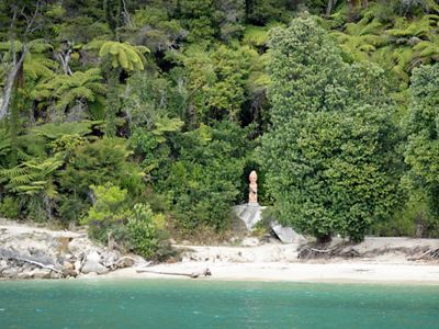 A pink statue stands in a niche within a dense forest at the edge of an ocean.