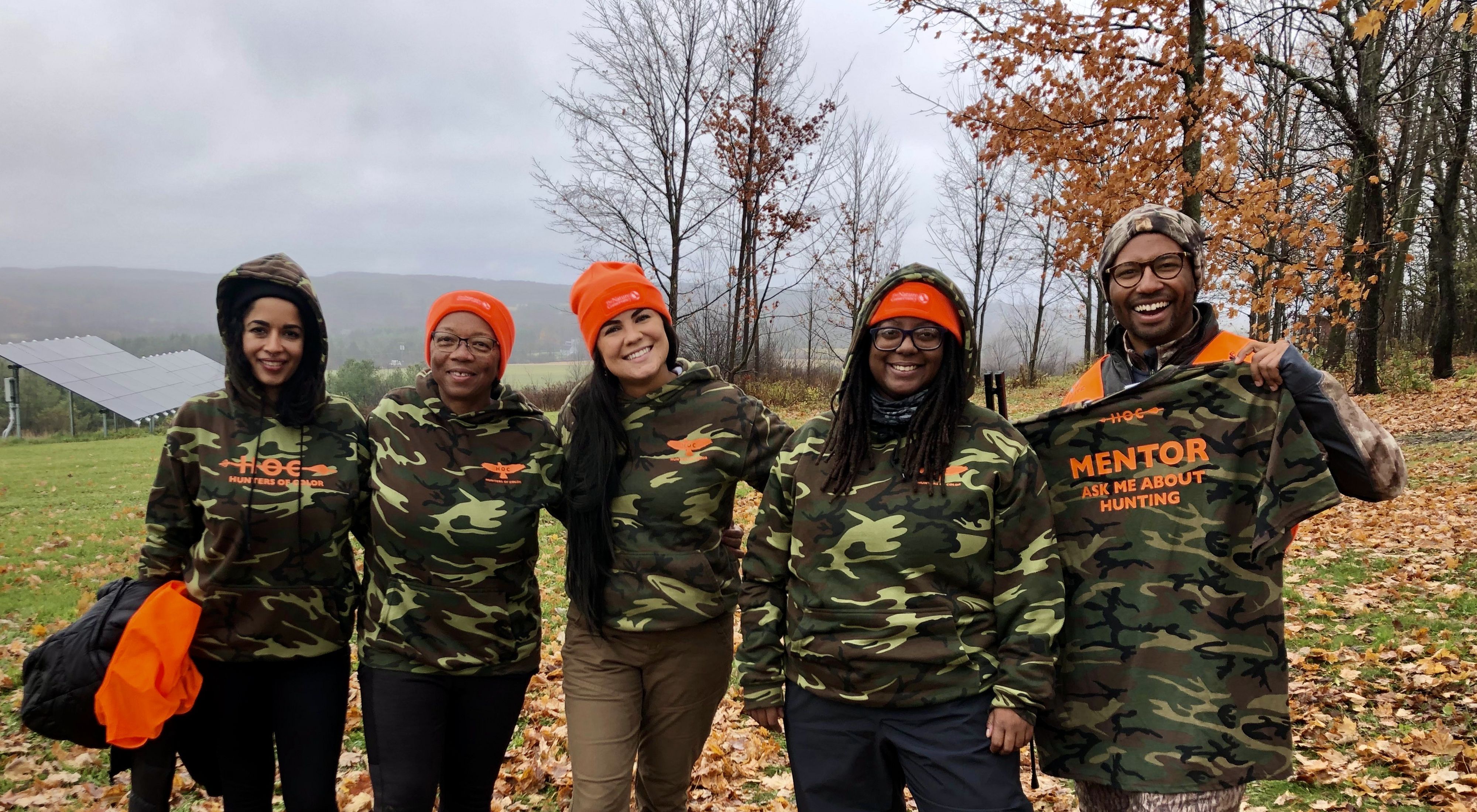 Five participants in the hunting weekend stand outdoors smiling at the camera. Each are wearing either a camouflaged t-shirt or sweatshirt that includes "HOC" printed.