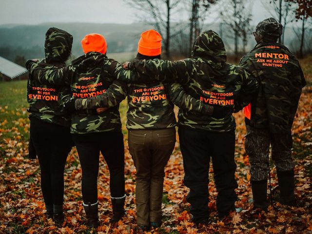 Five people stand with their backs turned to the camera. Each are wearing camouflage sweatshirts that contain writing in orange print. The writing says "The outdoors are for everyone," and "Mentor" 
