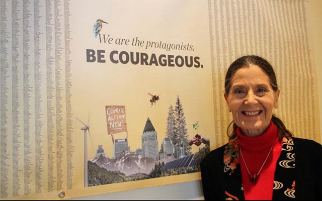 Lynn Scarlett stands in front of a poster that says, 'We are the protagonists. Be courageous', illustrated with buildings and nature and 25,000 signatures for climate action.