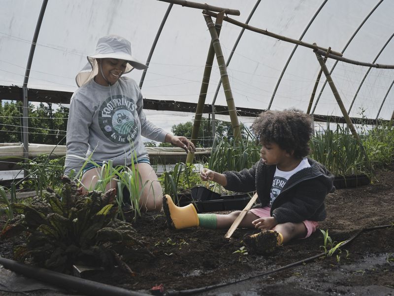 A woman and child kneel on the ground under the curved roof of an open-side greenhouse.