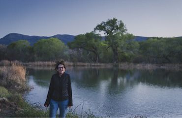 Una mujer sonríe frente a campos de cultivo, bosques y lago.