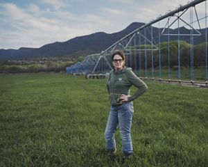 Claudia Hauser, wearing sunglasses, jeans and a green hoodie, stands with her hands on her hips in a verdant field of young grass, with her center pivot irrigation structure behind her.