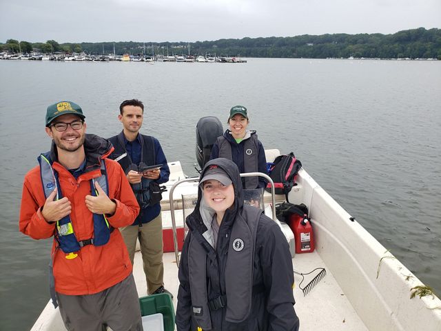Four crew members stand on a boat on Lake Ontario. The water is calm and the sky is grey as they prepare to leave. 