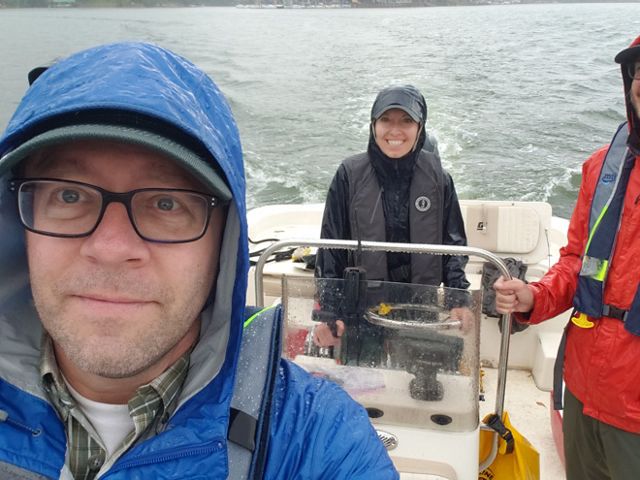 Three people stand on a boat in their rain gear as the rain falls. The surface of the water is slightly choppy.  