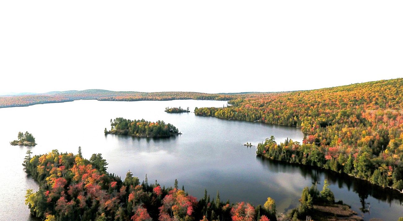 Aerial view of a lake surrounded by dense forest. The leaves display fall colors of red, orange and gold.