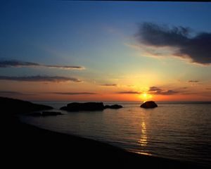 The sun sets over dark, calm waters, while clouds float in the darkening sky overhead.