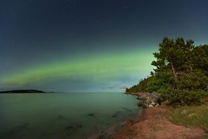 Green Aurora Borealis stretch over the horizon at night. In the foreground is a rocky, coniferous Upper Peninsula Coastline against calm Lake Superior.