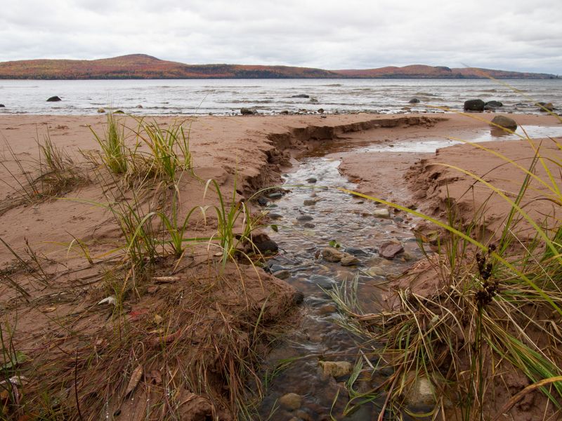 A stream of water runs through a sandy and rocky area and into a body of water in the Keweenaw Peninsula.