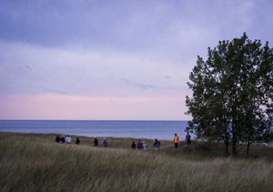 A line of people in the distance hike through tall dune grasses, with Lake Michigan visible just beyond. The sky is a dusky pink and purple, and there is a single tree in the frame.