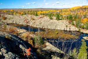 A forest of trees in the autumn and rocky cliffs surround a dark body of water.  
