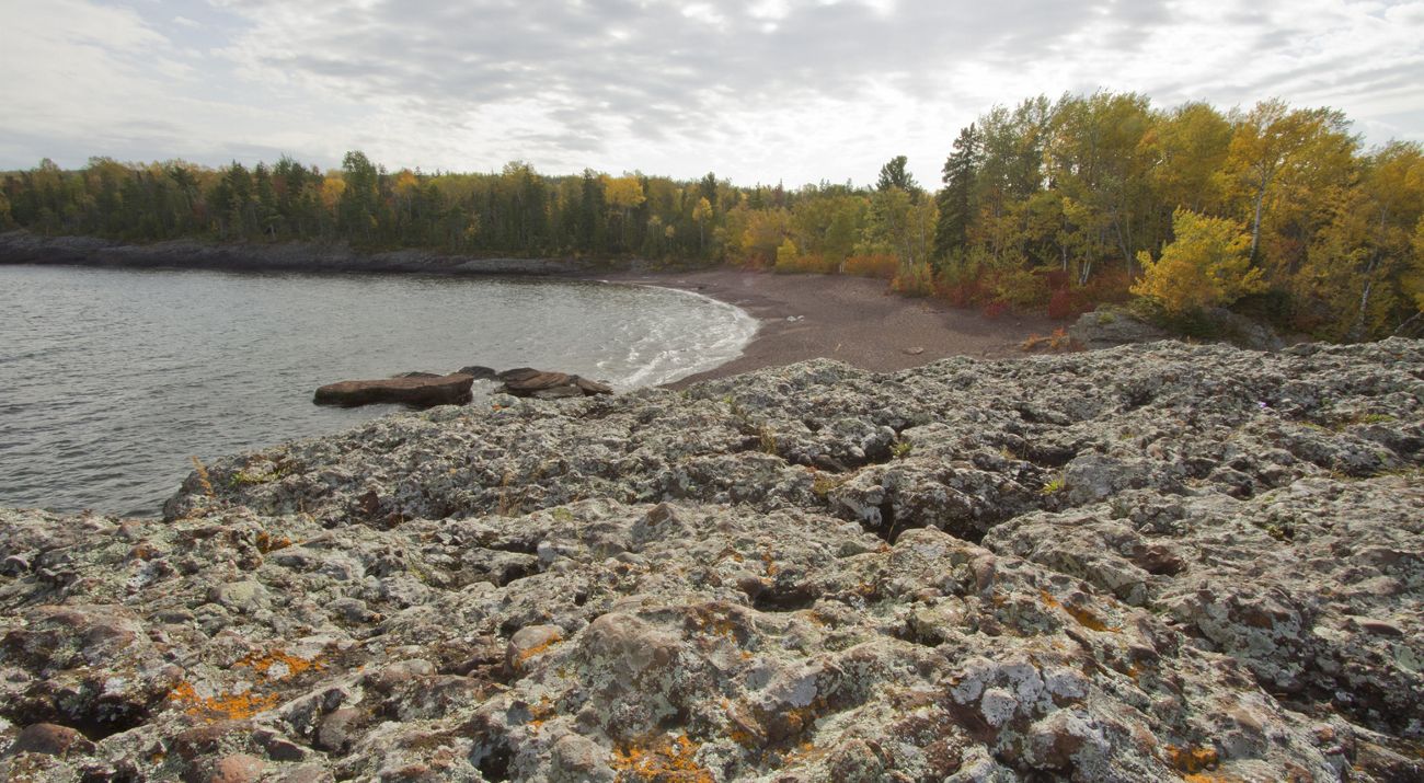 A rocky outcropping overlooks a rocky beach and fall-colored trees.