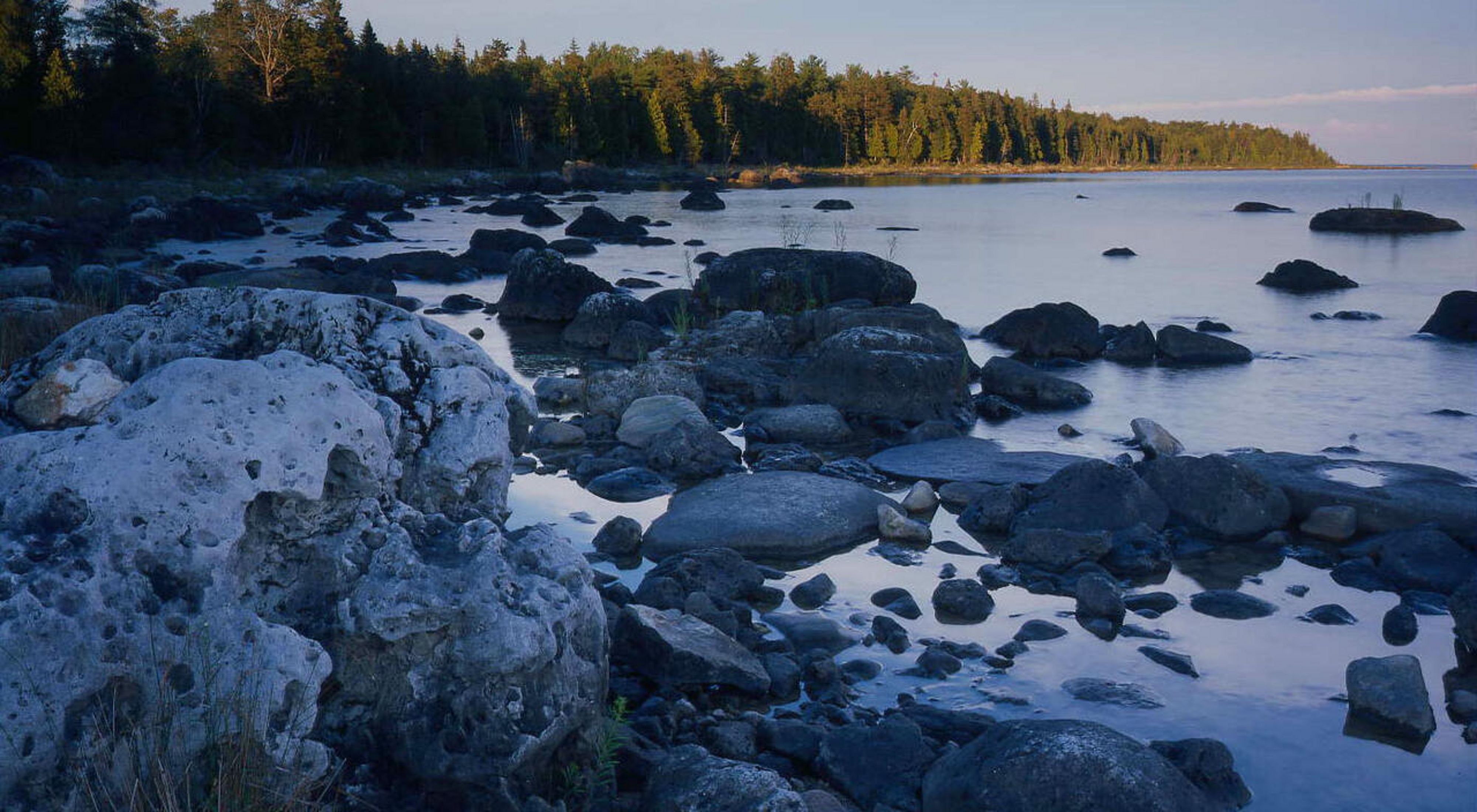 Rocks of various sizes along the shore of a lake. A forest grows on the land surrounding the lake. 