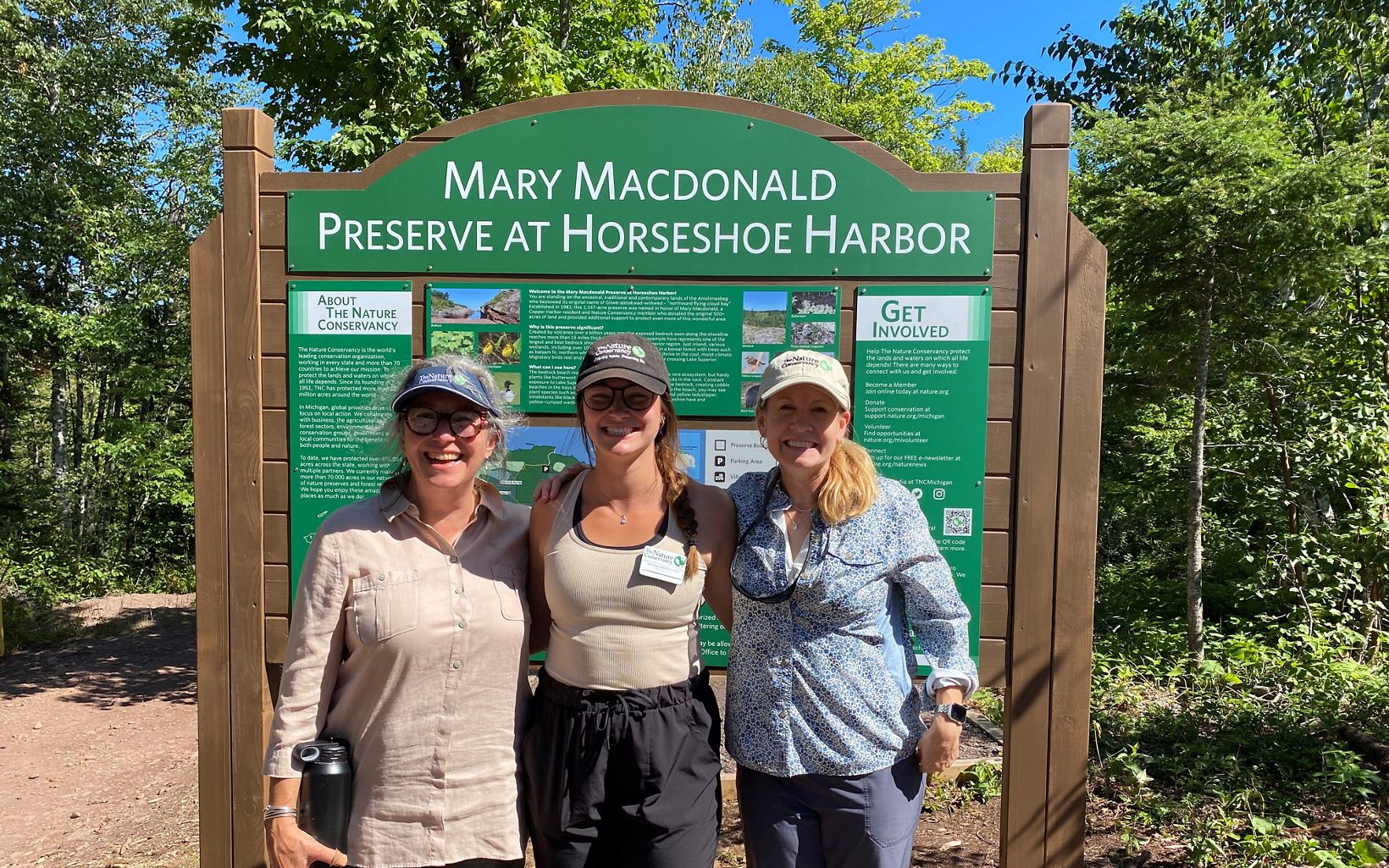 Three people pose for a photo in front of a preserve kiosk sign on a sunny day. 