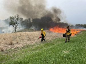 Two people in protective gear walk along a preserve as the manage a prescribed fire burning in the background.