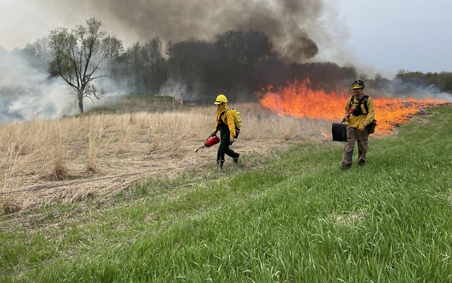 Two people in protective gear walk along a preserve as the manage a prescribed fire burning in the background.