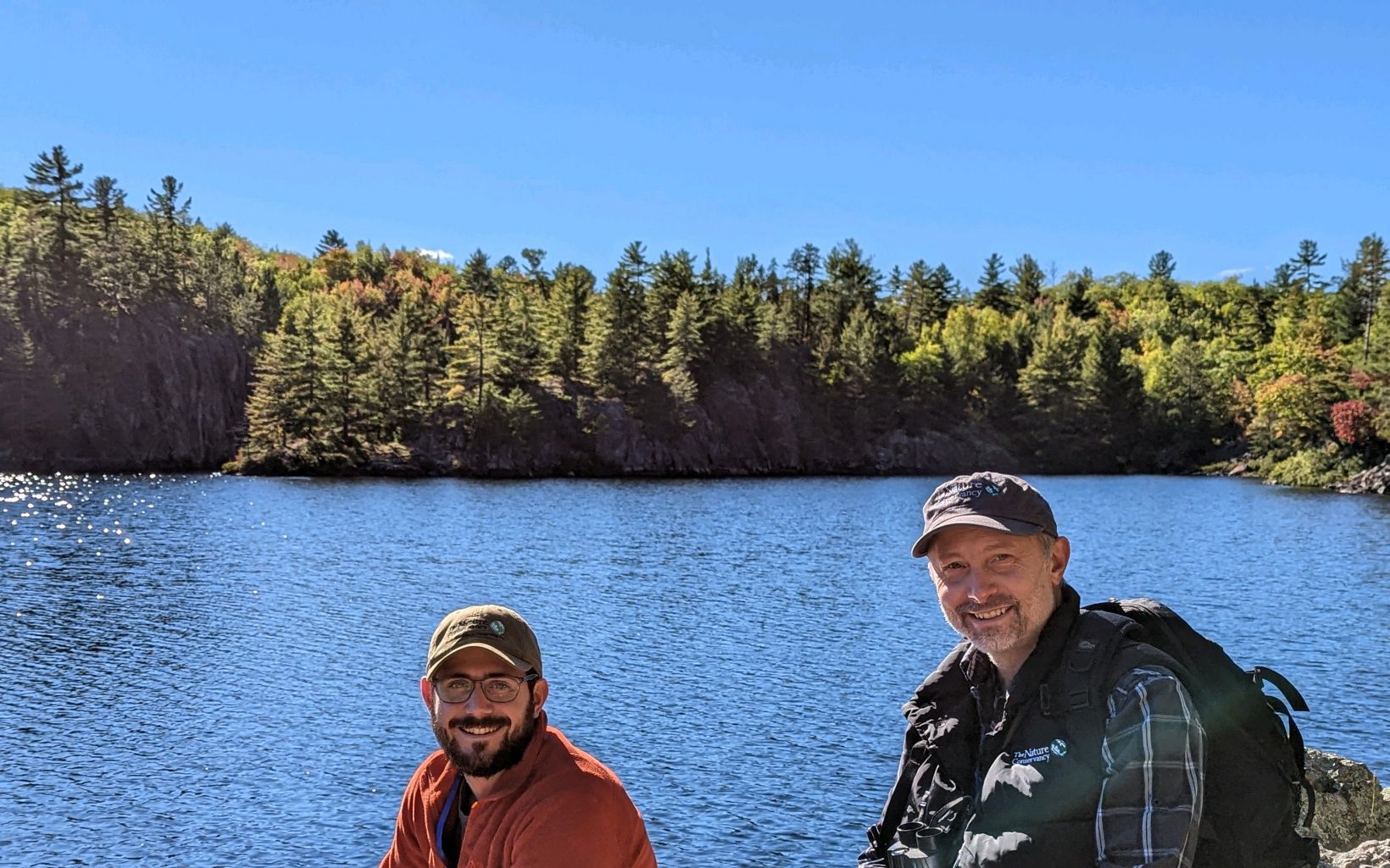Two staff members smiling with a lake and forested shoreline in the distance. 
