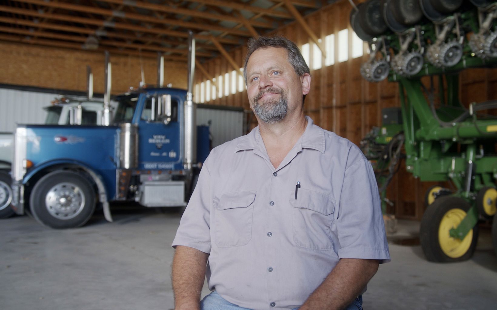 Scott Brechtelsbauer sits in a barn and is surrounded by farm equipment.