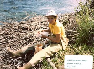 A woman sits on a bed of sticks at the edge of a body of water.