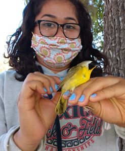 Young woman in with dark-framed eyeglasses and colorful face mask holds bright yellow bird.