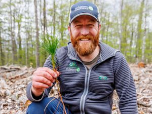 A man smiling and wearing a gray jacket with The Nature Conservancy logo on it is crouching down and holding a tree seedling.