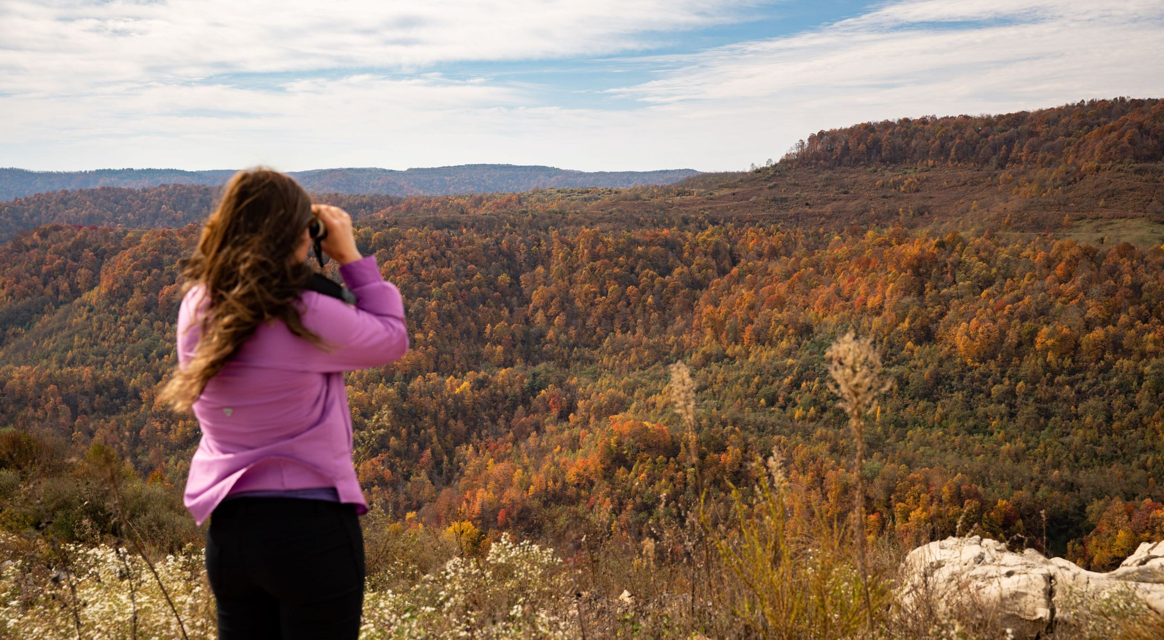 A woman looks at a colorful fall Appalachian forest through binoculars.