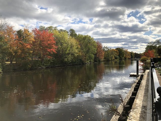 The Taunton River, reflecting the gray sky, has autumnal trees lining it on one side and a waterfront walkway on the other side.