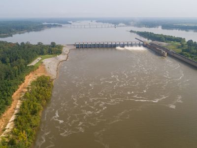 River surrounded by woods with a lock and dam and a bridge in the background.