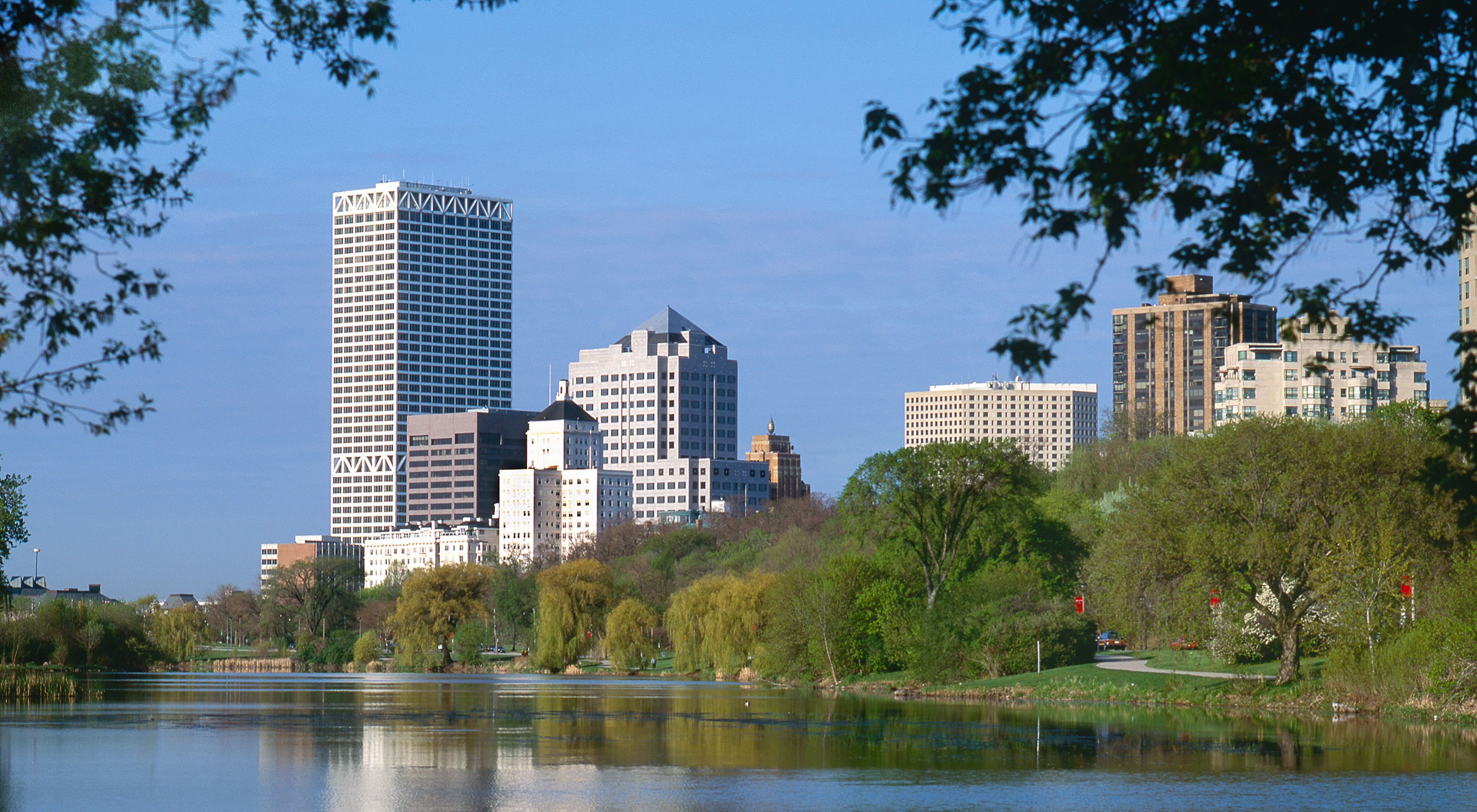 View of a city skyline with tall buildings in the background and trees and grass along the shoreline of a small lake.