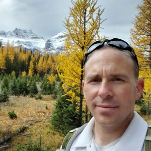 Person smiling in front of trees and mountains.