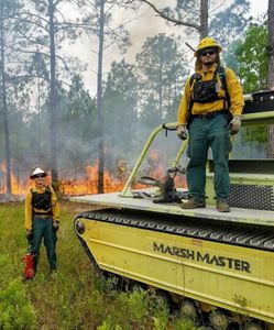 Two men wearing yellow clothing and hard hats pose in front of a forest that is being burned.