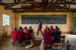 A man stands at the front of a classroom, and a group of students in red clothing look up at him and a chalkboard.
