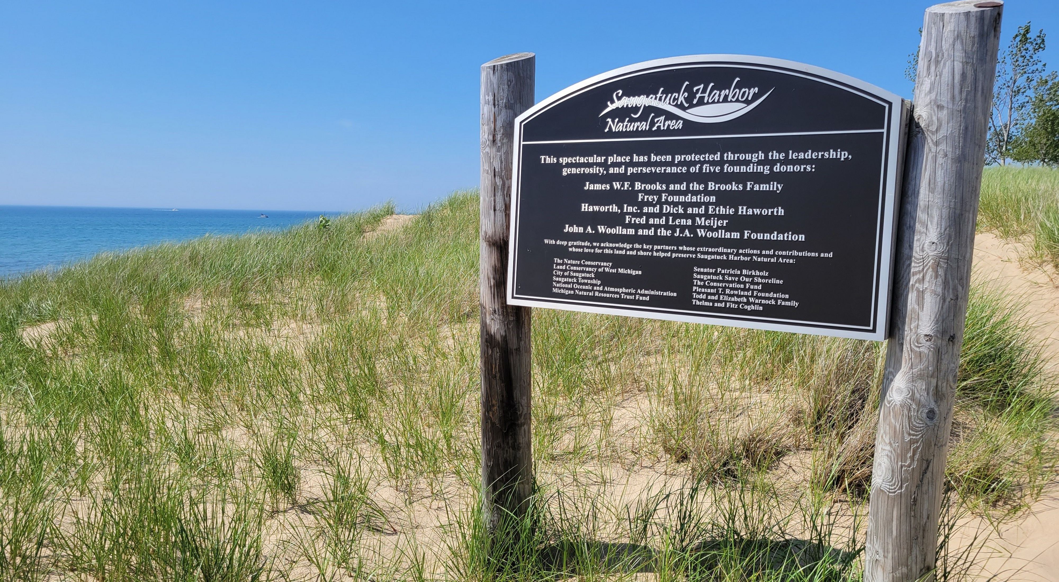 A sign for Saugatuck Harbor Natural Area stands in a grassy, sandy area. 