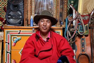 a man in a red coat and brown hat with a saddle and other equipment behind him.