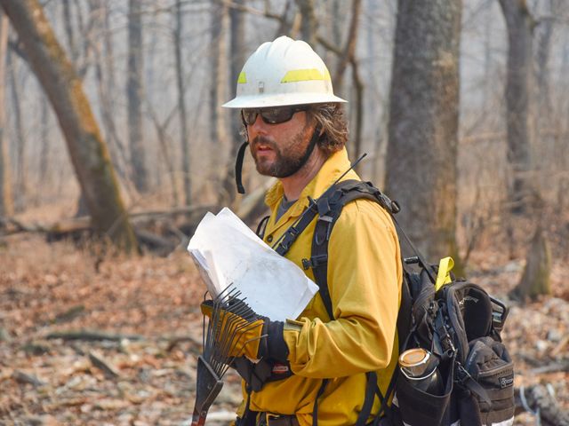 Steve Woods in full fire yellow fire gear stands in a forested landscape holding papers and a rake.