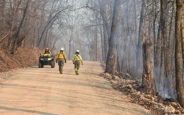 Two fire fighters walk along a rural forested road as a women drives by on a UTV.