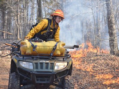 Megan Alkazoff in yellow fire gear drives a UTV along a rural dirt road with a line of fire to the right of her.