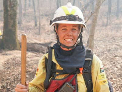 Kylie Paul in full yellow fire gear smiles at the camera with a forested landscape behind her.