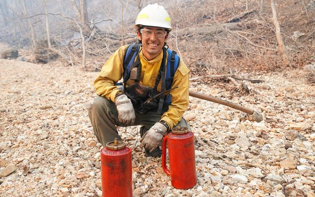 Eric Chien in a yellow fire jacket and white helmet kneels in a rocky creek bed with two red drip torches. 