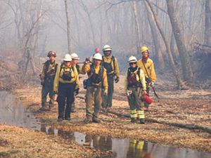 A small group of fire fighters walk along a creek bed in a forested area with smoke from a prescribed burn behind them.
