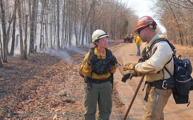 Autumn Jensen and Ryan Gauger in fire gear talk along a rural dirt road with smoke from a prescribed fire behind them.